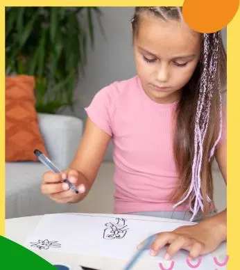 Young girl with braided hair in a pink shirt concentrating while coloring a picture with markers at a table in a cozy indoor setting.